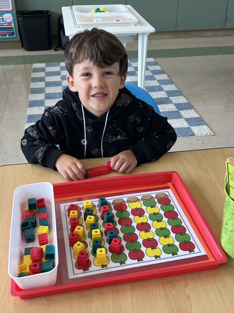 Young boy smiling while playing a color-matching game with red, yellow, and green blocks on a tray at a classroom table.
