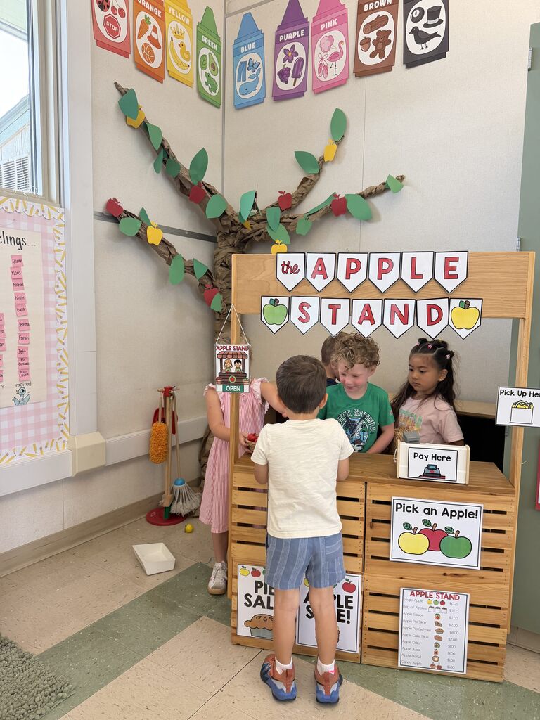 Children role-play buying and selling apples at a classroom apple stand decorated with a paper apple tree.