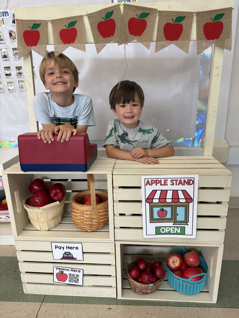 Two smiling children stand behind a pretend apple stand stocked with baskets of red apples and a toy register.