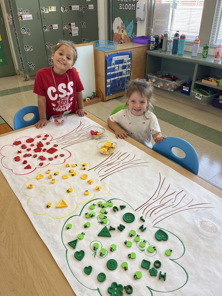 Two children smile beside a table while sorting red, yellow, and green manipulatives into matching tree outlines.