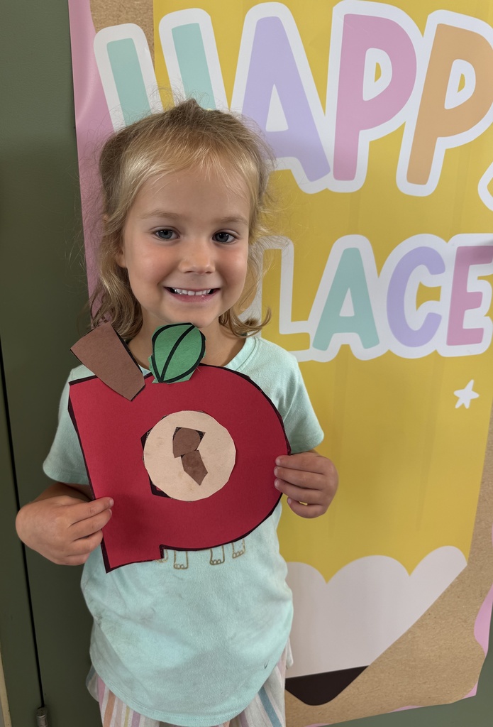 A smiling child holds a handmade red paper apple craft in front of a colorful classroom display.