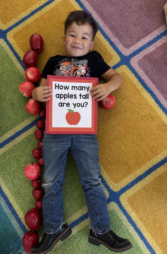 A smiling child lies beside a row of apples used to measure height while holding a “How many apples tall are you?” sign.