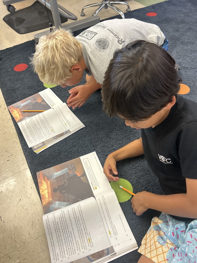 Two young boys sitting on a carpet reading and discussing open books with pencils in hand in a classroom setting.