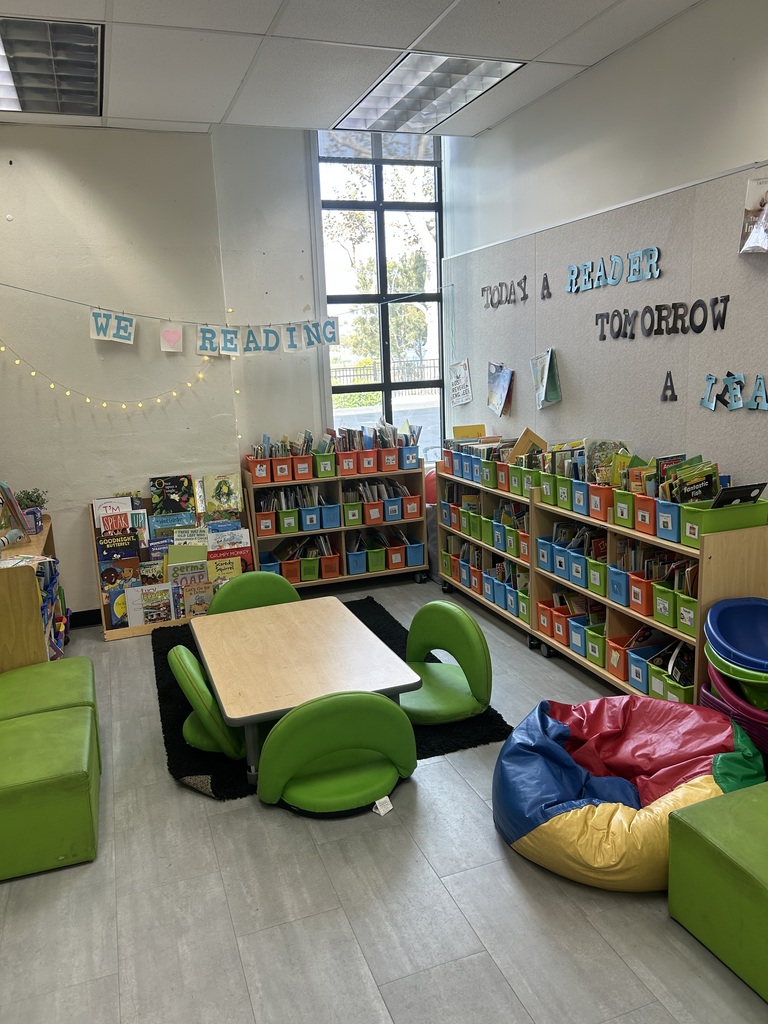 Cozy classroom reading corner with colorful bookshelves, green floor chairs, and a bean bag under a large window.