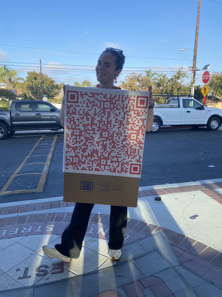 Person standing outdoors wearing a large cardboard QR code costume on a sunny day with parked cars in the background.