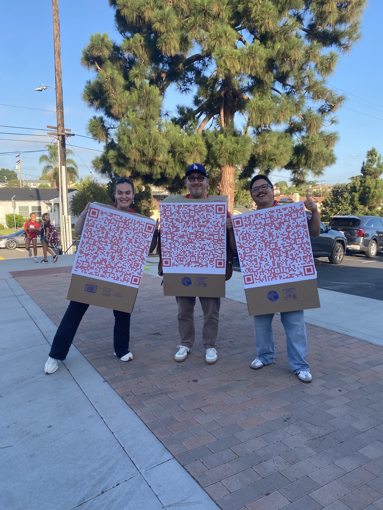 Three people standing outdoors holding large QR code posters in front of them on a sunny day.
