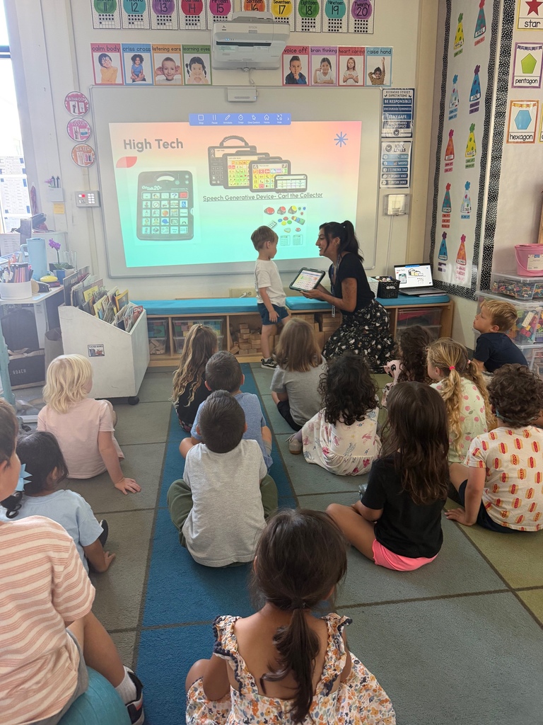A teacher demonstrates a speech-generating communication device with a student as classmates watch attentively from the classroom rug.