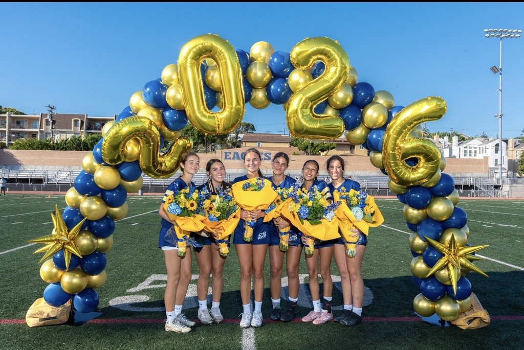 Six female athletes in blue uniforms holding bouquets stand under a gold and blue balloon arch shaped as 2026 on a sports field.