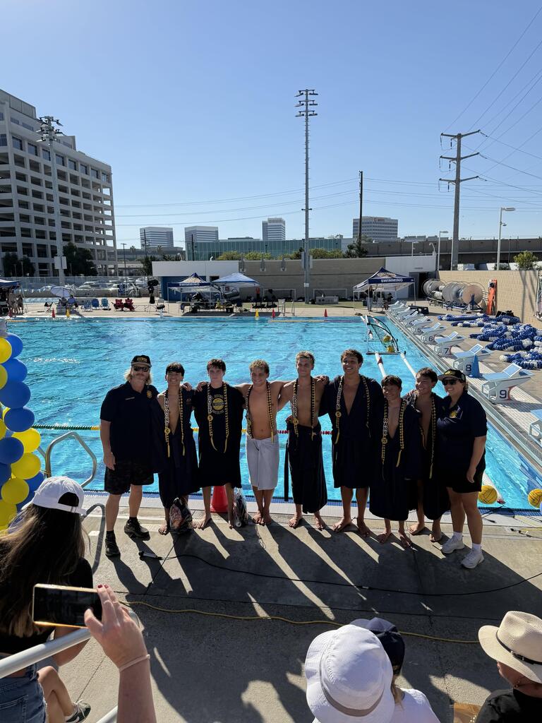 Seven senior swimmers pose with two coaches beside an outdoor pool, wearing black and gold leis for Senior Recognition.