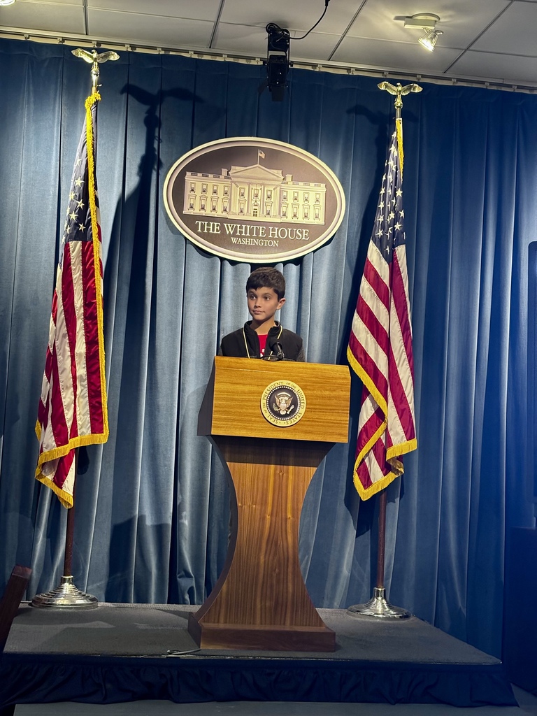 A student stands at a White House press briefing podium between two American flags, beneath the White House emblem.
