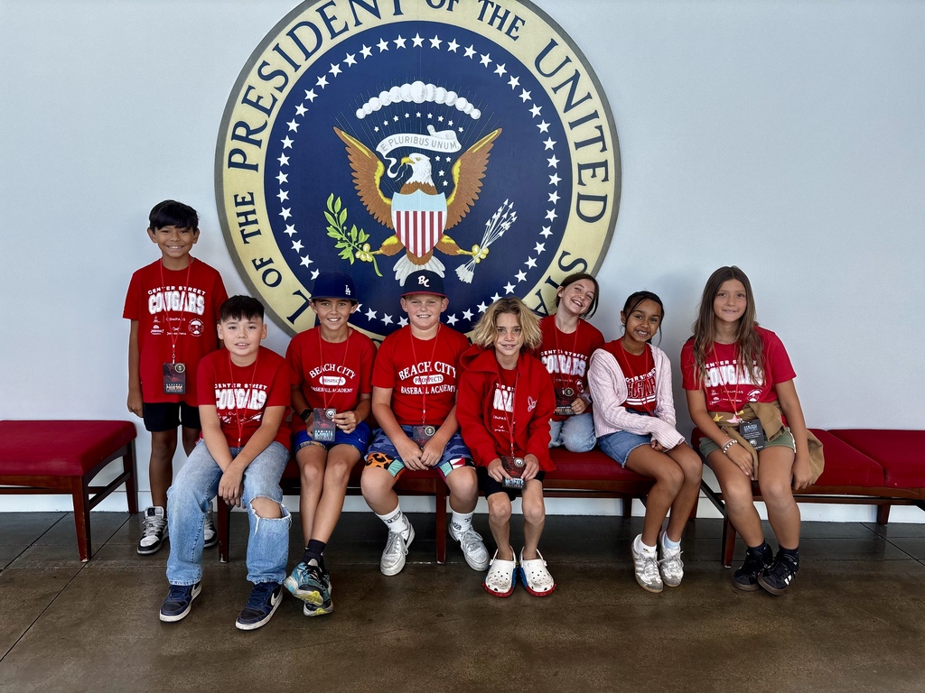 Eight smiling students in red shirts pose together beneath a large presidential seal during their educational tour.