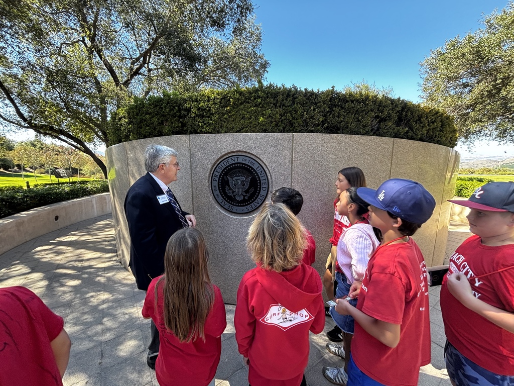 A guide speaks with students in red shirts beside the presidential seal displayed on a stone wall outdoors.