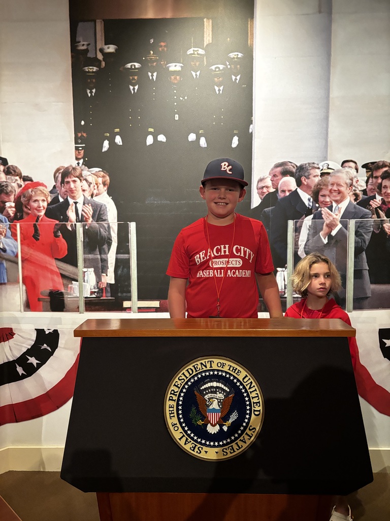 Two students pose behind a presidential podium with the official seal and a historic inauguration photograph in the background.