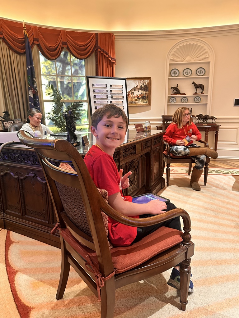A smiling student flashes a peace sign while seated in a replica Oval Office with an ornate desk and furnishings.