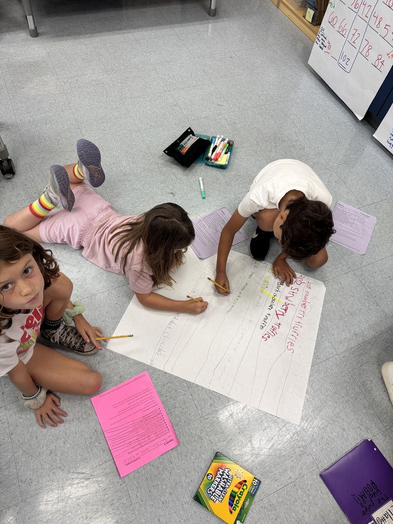 Three children working together on a large poster with markers and pencils on a classroom floor.