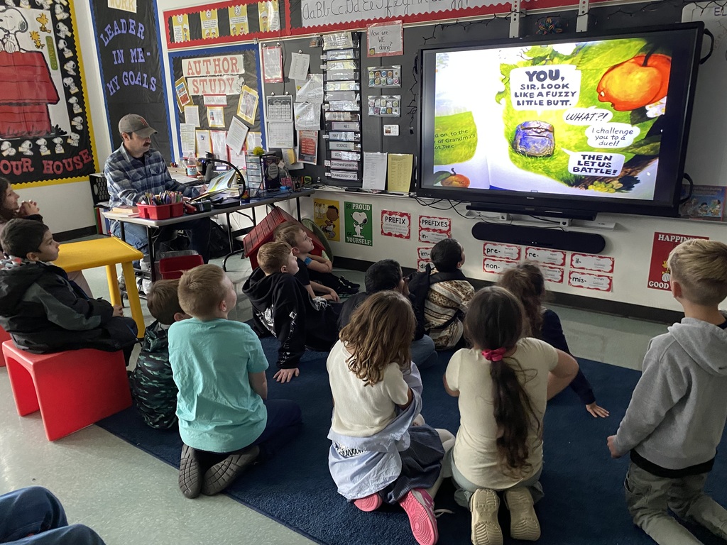 Image of a 4th grade class sitting in front of a smartboard looking at a story that is being projected from a document camera
