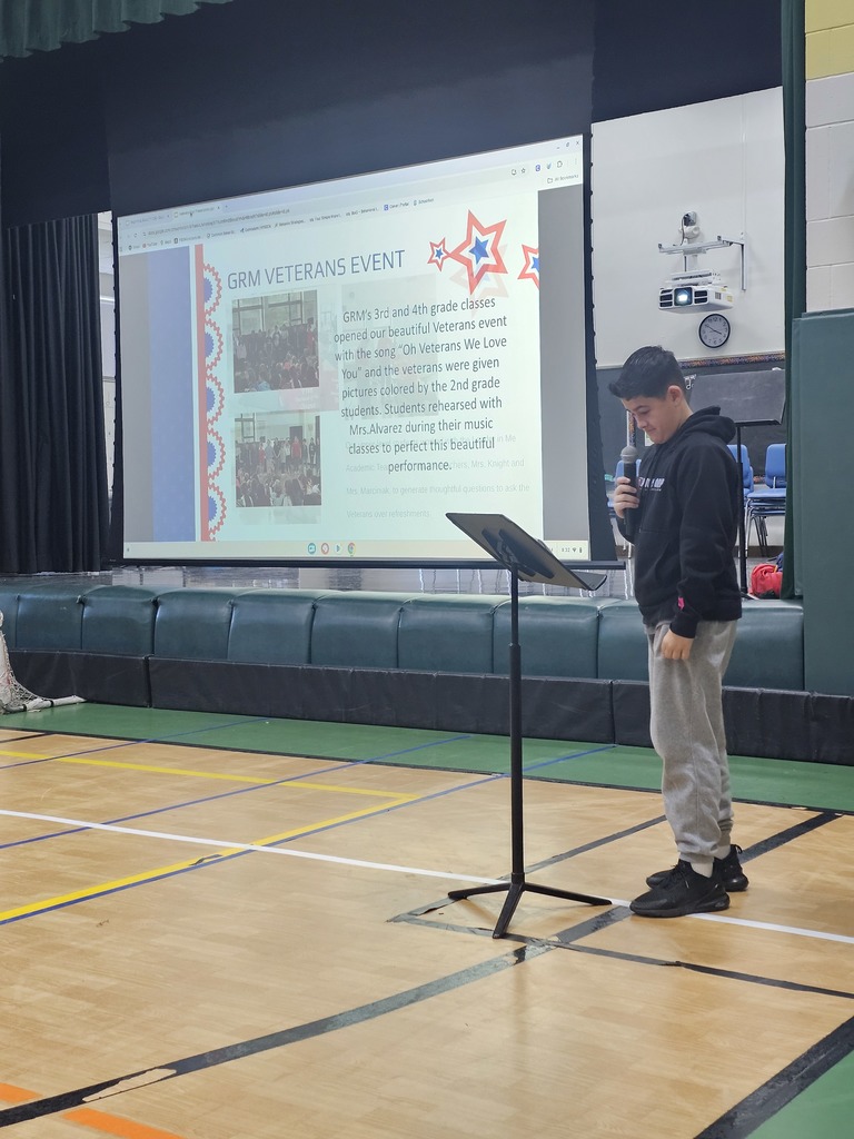 Image of a student standing at a microphone in front of a screen that lays out a Veterans Day event