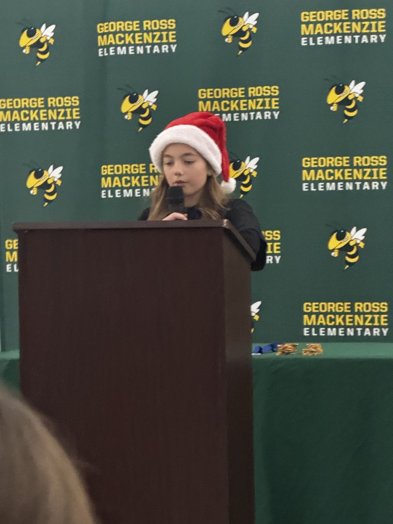 Image of a student wearing a Santa hat, standing at a podium in front of a green & yellow background with the George Ross Mackenzie Elementary yellowjacket logo