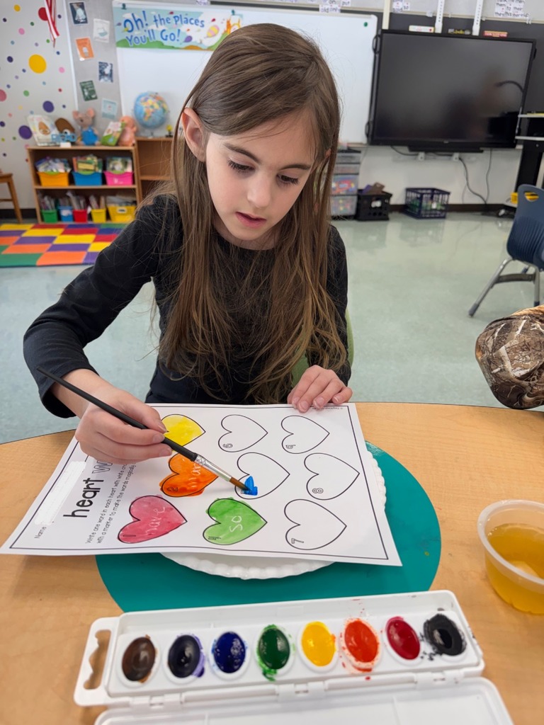 A student is seen with a paintbrush painting their "heart" words worksheet