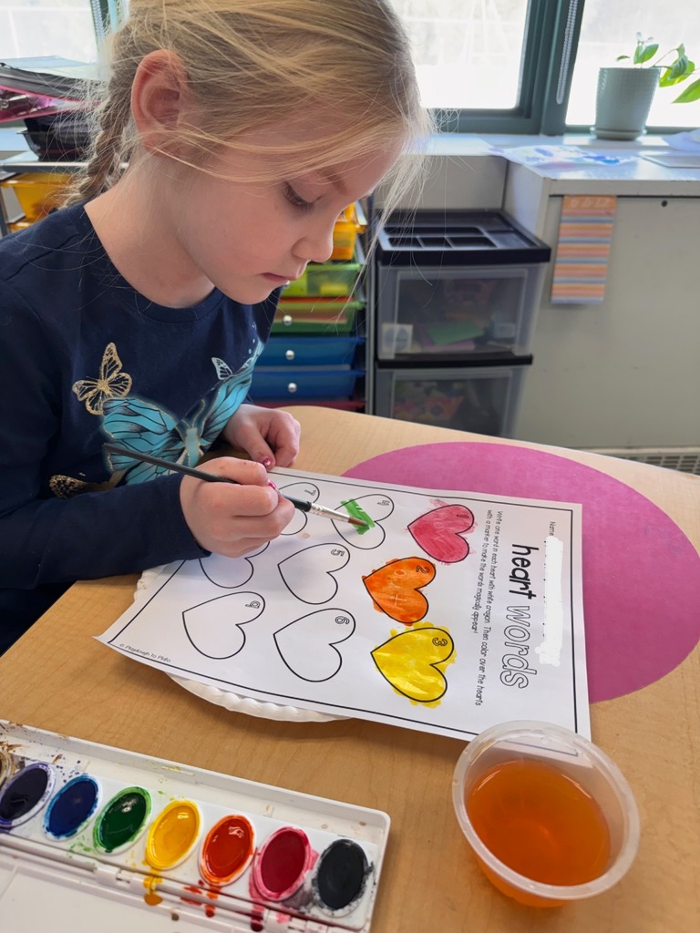 A student is seen with a paintbrush painting their "heart" words worksheet