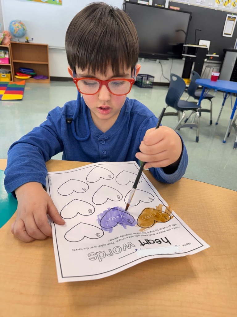 A student is seen with a paintbrush painting their "heart" words worksheet