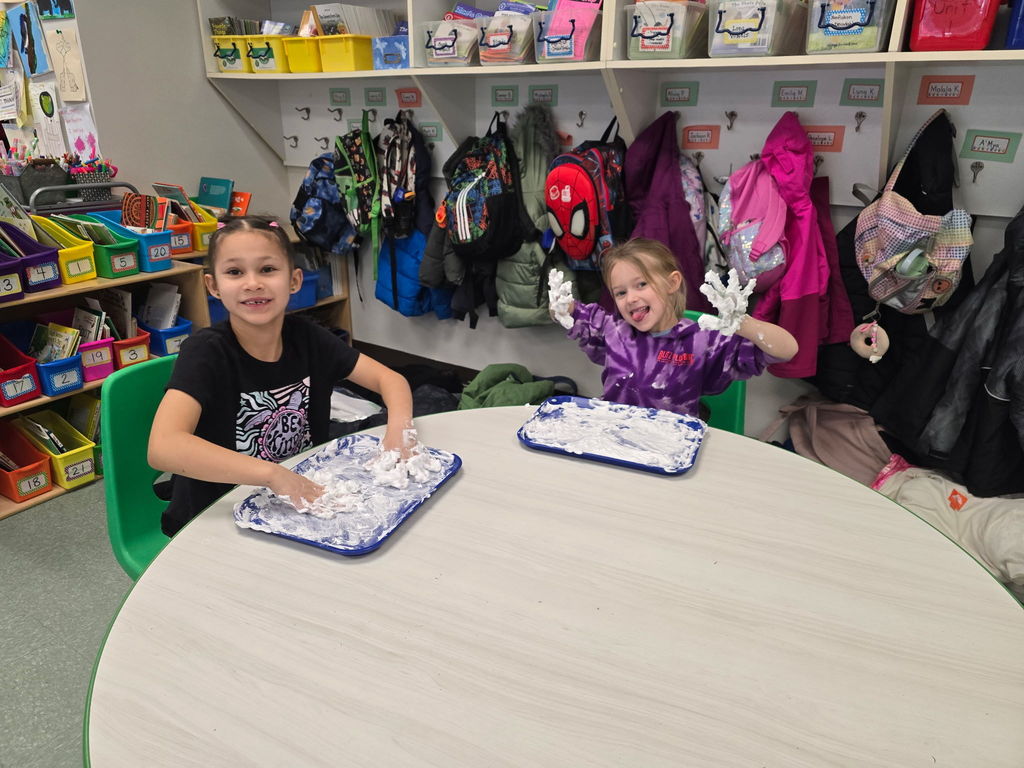 Students sit at round tables with a tray of shaving cream they are using to practice their phonics skills and pose for the camera with saving cream on their hands