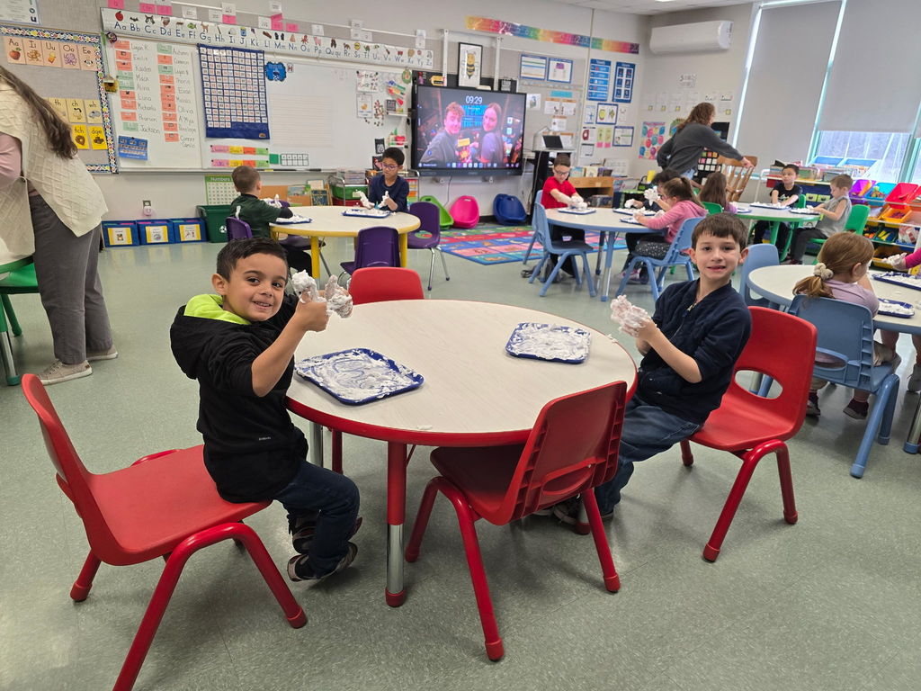 Students sit at round tables with a tray of shaving cream they are using to practice their phonics skills and pose for the camera with saving cream on their hands