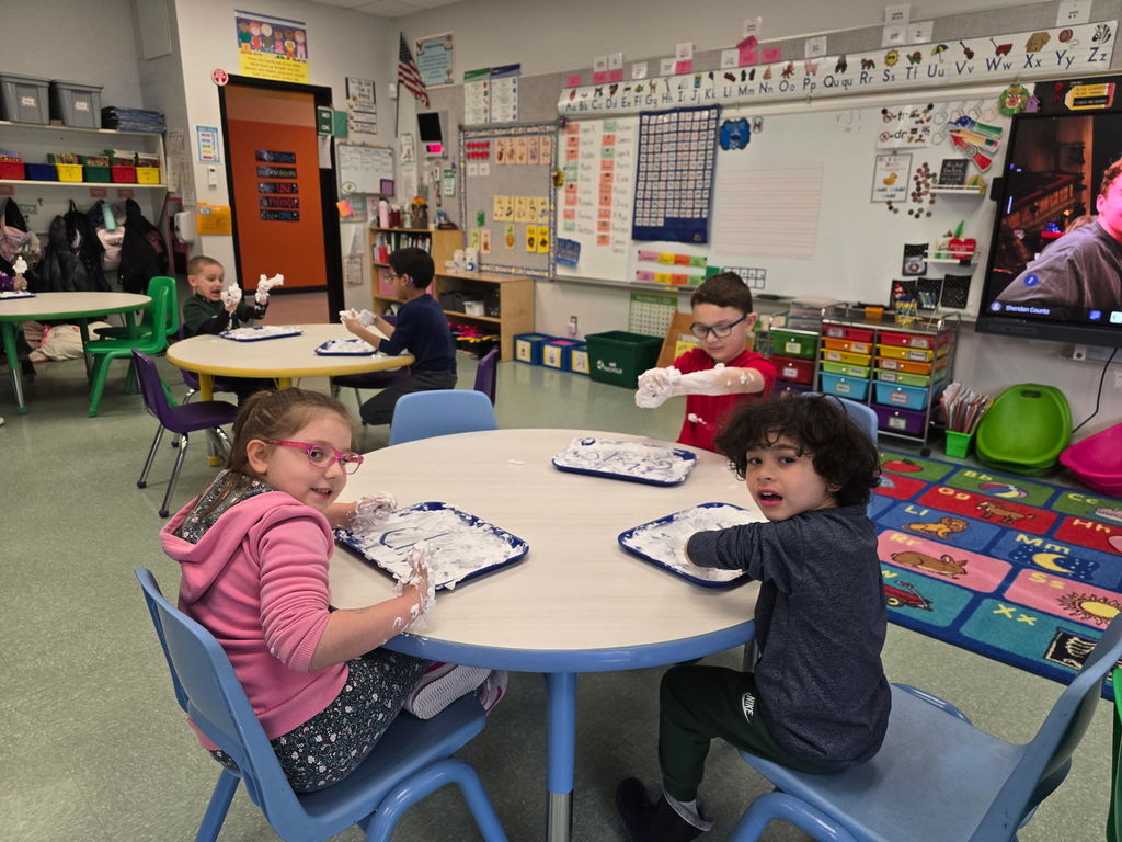 Students sit at round tables with a tray of shaving cream they are using to practice their phonics skills and pose for the camera with saving cream on their hands