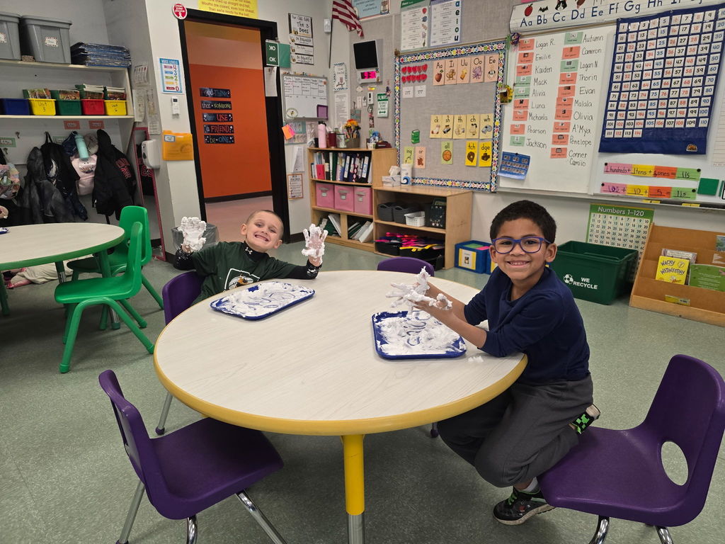 Students sit at round tables with a tray of shaving cream they are using to practice their phonics skills and pose for the camera with saving cream on their hands