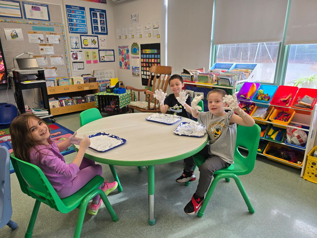 Students sit at round tables with a tray of shaving cream they are using to practice their phonics skills and pose for the camera with saving cream on their hands