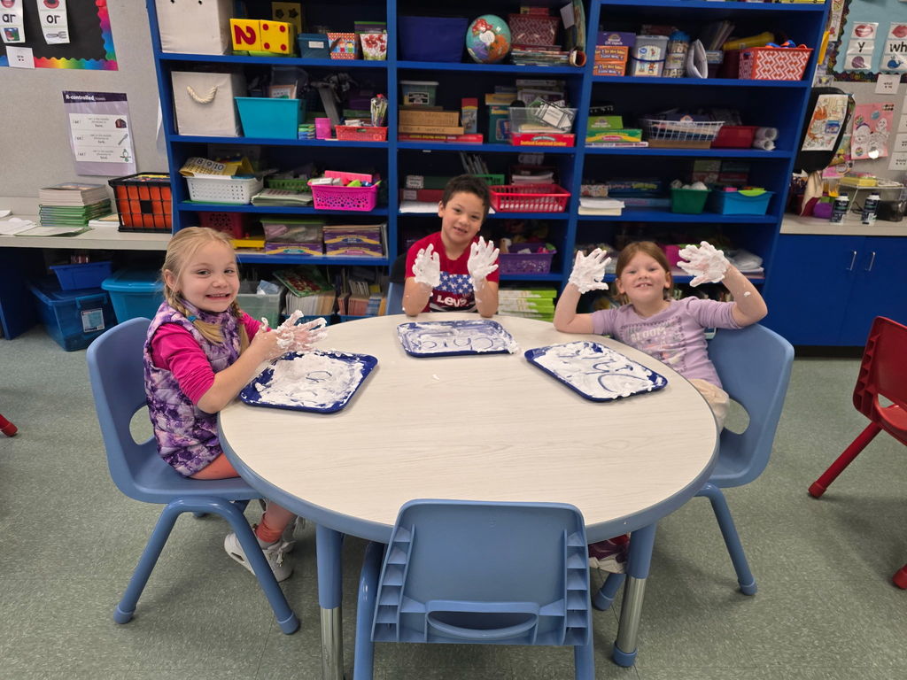 Students sit at round tables with a tray of shaving cream they are using to practice their phonics skills and pose for the camera with saving cream on their hands