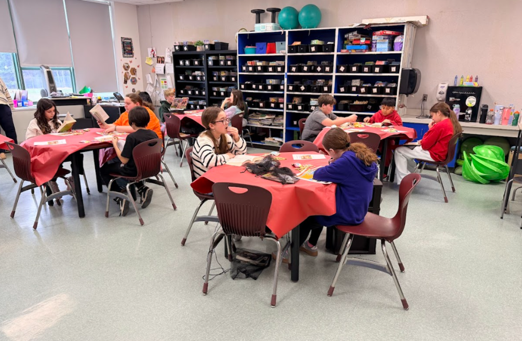 Image of children sitting in a classroom at round tables with red table cloths reading books and eating snacks
