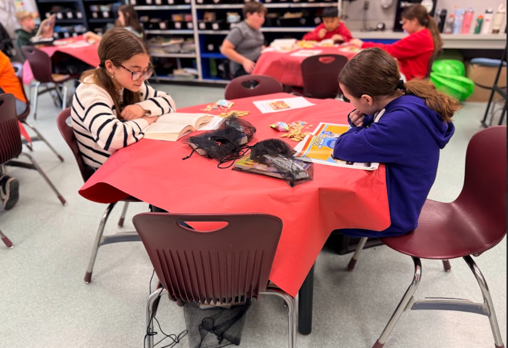 Image of children sitting in a classroom at round tables with red table cloths reading books and eating snacks