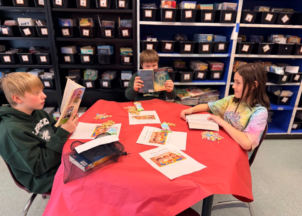 Image of children sitting in a classroom at round tables with red table cloths reading books and eating snacks