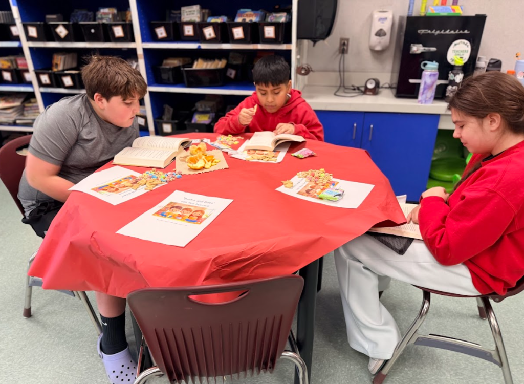 Image of children sitting in a classroom at round tables with red table cloths reading books and eating snacks