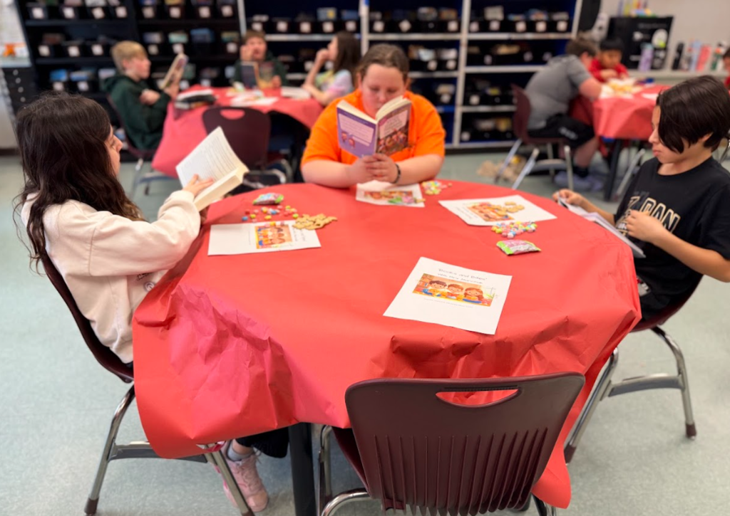 Image of children sitting in a classroom at round tables with red table cloths reading books and eating snacks