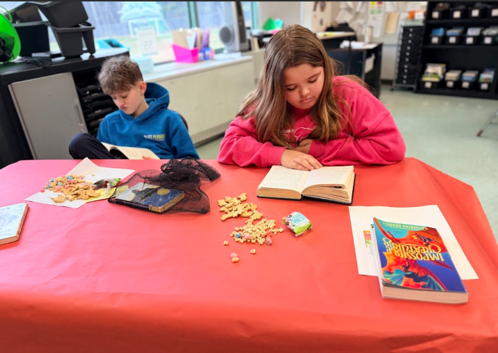 Image of children sitting in a classroom at round tables with red table cloths reading books and eating snacks