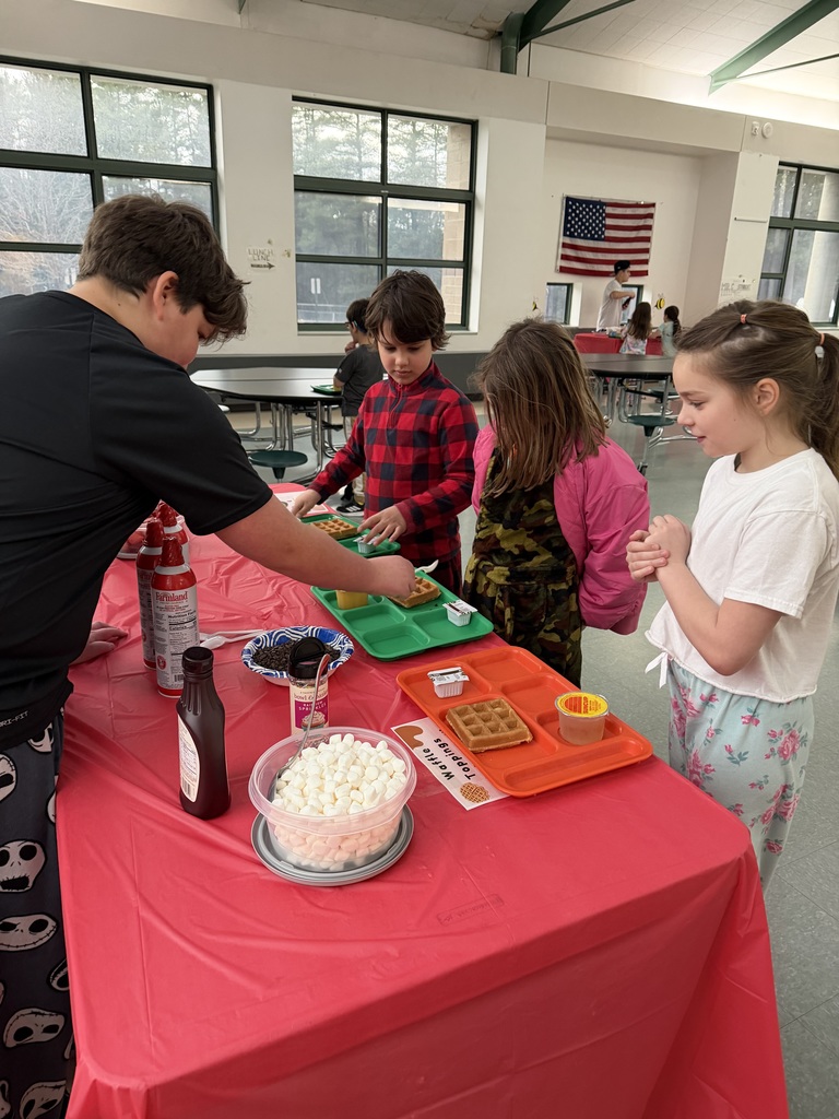 A  students stands at a table covered with a red table cloth as he helps younger students put toppings on a waffle