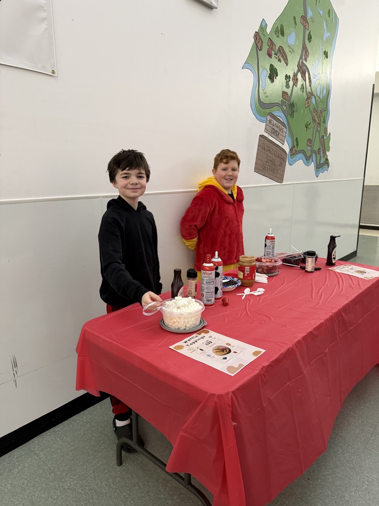 Two students stand in front of a table covered with a red table cloth. The table has fixings for a waffle toppings bar including marshmallows, fresh berries, whipped cream, syrup and sprinkles