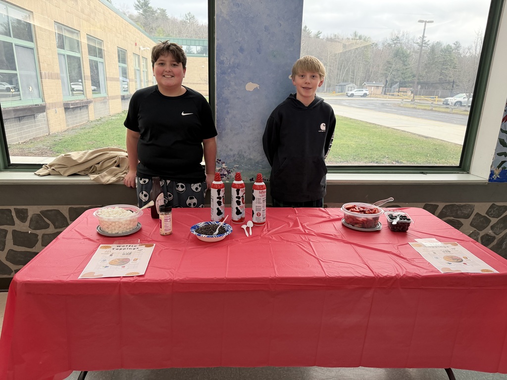 Two students stand in front of a table covered with a red table cloth. The table has fixings for a waffle toppings bar including marshmallows, fresh berries, whipped cream, syrup and sprinkles