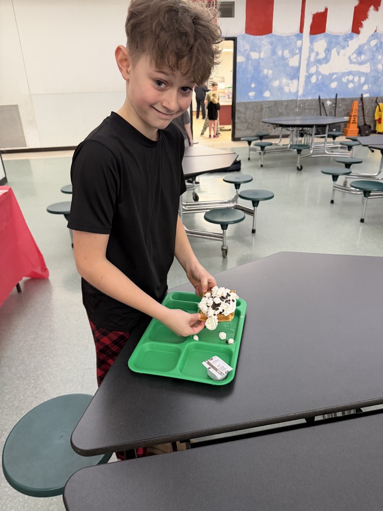 A student stands  at a cafeteria table with a green tray holding a waffle topped with what looks like marshmallows and chocolate chips.