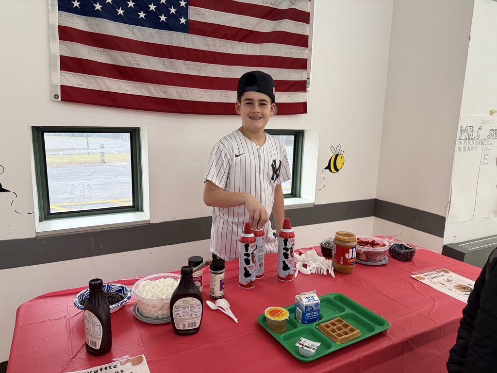 A student stands in front of a table covered with a red table cloth. The table has fixings for a waffle toppings bar including marshmallows, fresh berries, whipped cream, syrup and sprinkles
