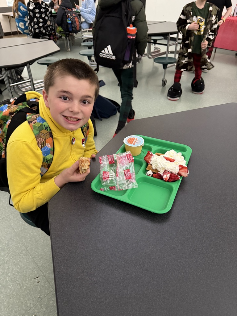 A student sits at a cafeteria table with a green tray that has a waffle with strawberries & whipped cream