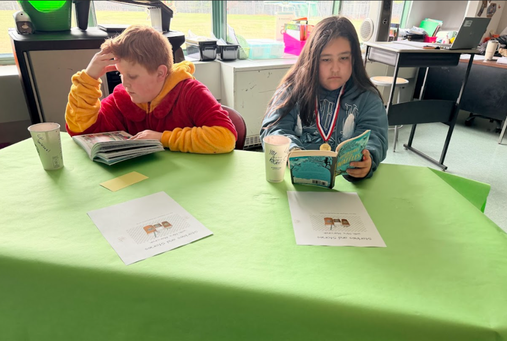Image of student sitting around a table with a green table cloth, enjoying hot cocoa while reading a book
