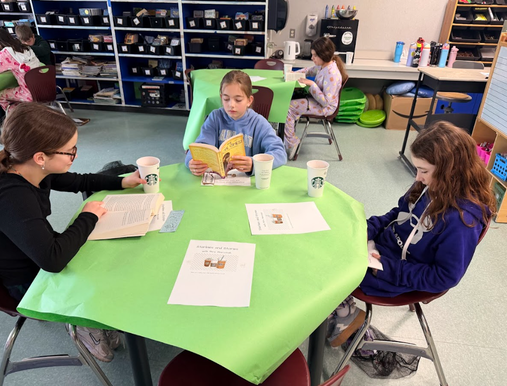 Image of student sitting around a table with a green table cloth, enjoying hot cocoa while reading a book