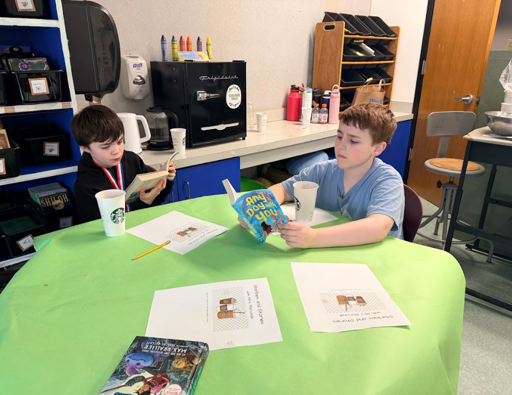 Image of student sitting around a table with a green table cloth, enjoying hot cocoa while reading a book