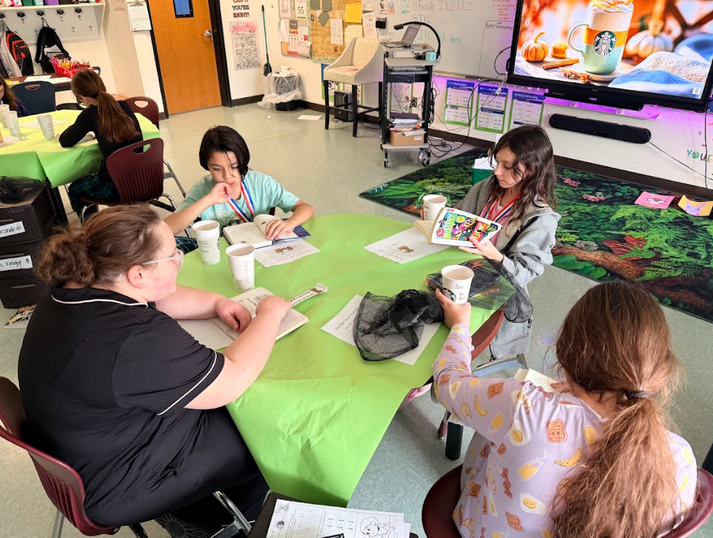 Image of student sitting around a table with a green table cloth, enjoying hot cocoa while reading a book