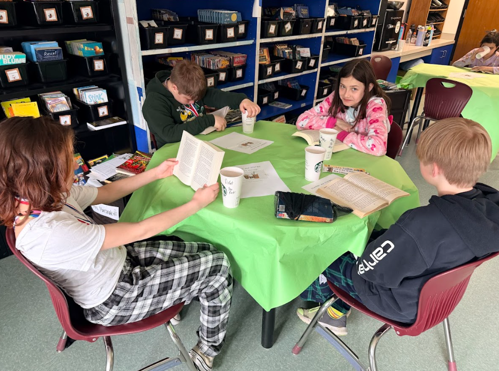 Image of student sitting around a table with a green table cloth, enjoying hot cocoa while reading a book