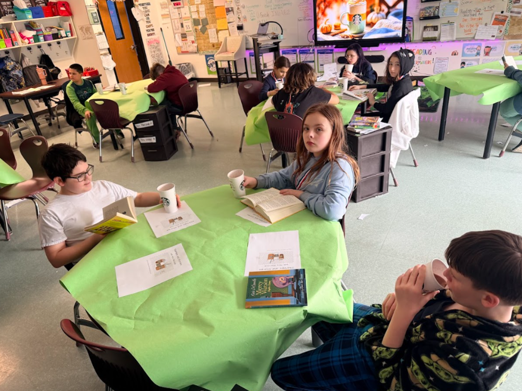 Image of student sitting around a table with a green table cloth, enjoying hot cocoa while reading a book