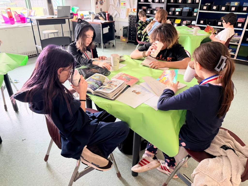 Image of student sitting around a table with a green table cloth, enjoying hot cocoa while reading a book
